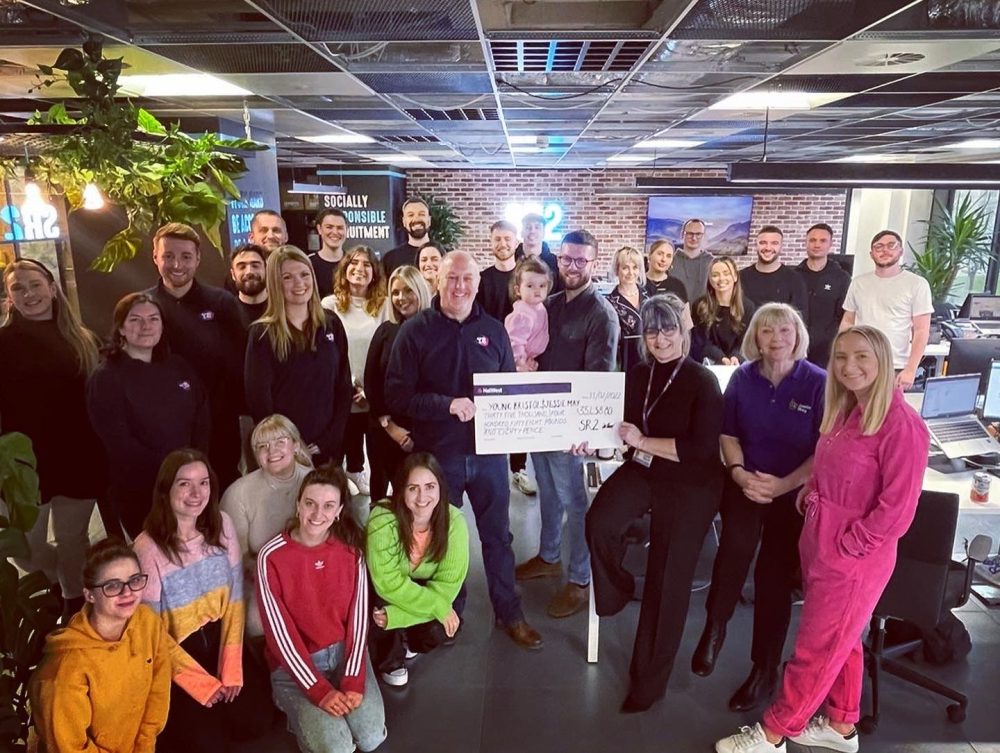Group of individuals in an office room crowded round a giant cheque
