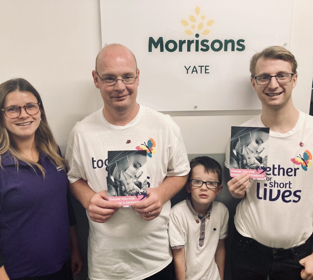 3 adults, one woman wearing purple Jessie May polo shirt, and 2 men wearing white Together for Short Lives t-shirts holding cards, with young boy wearing white polo shirt