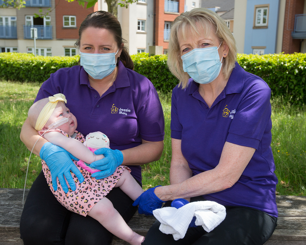 Two Jessie May nurses, female, wearing purple polo shirts, surgical masks and gloves holding a young child, girl, with a nasal-tube